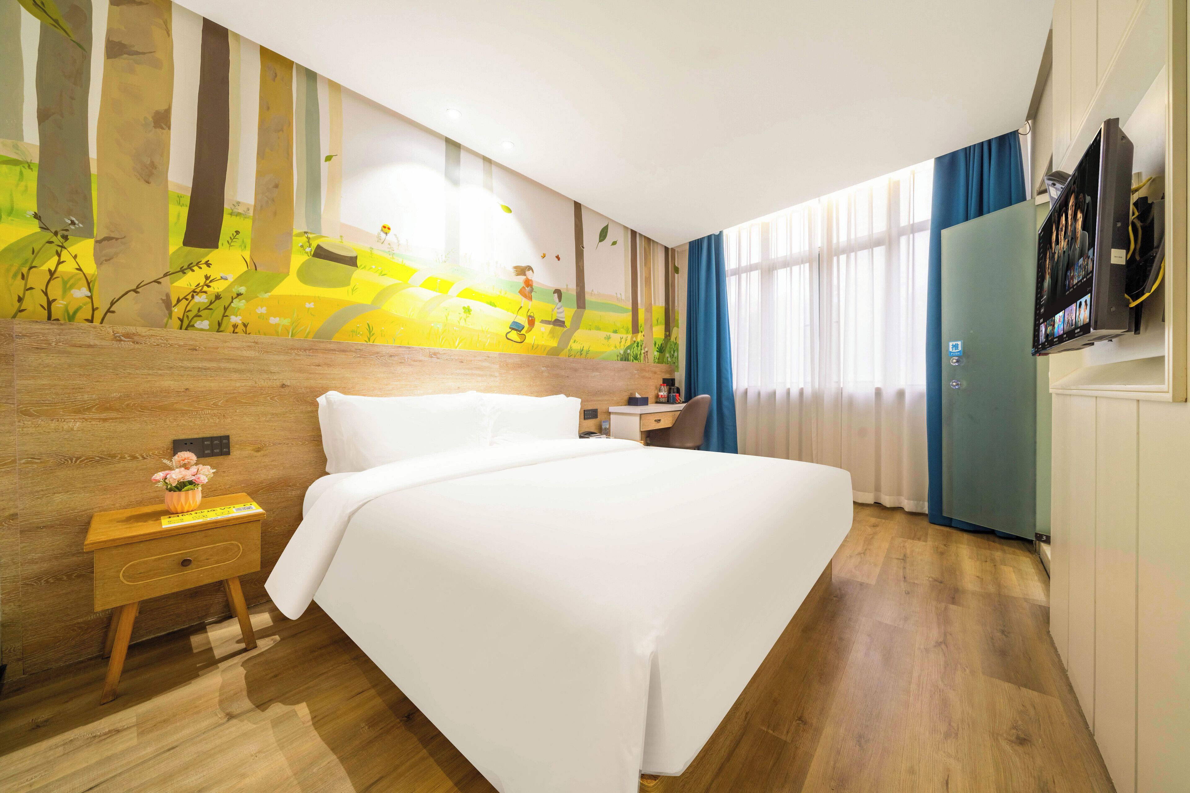 A hotel room with a large bed, wooden headboard, a nightstand with a vase of flowers, a desk with a chair, a TV mounted on the wall, and a window with curtains.