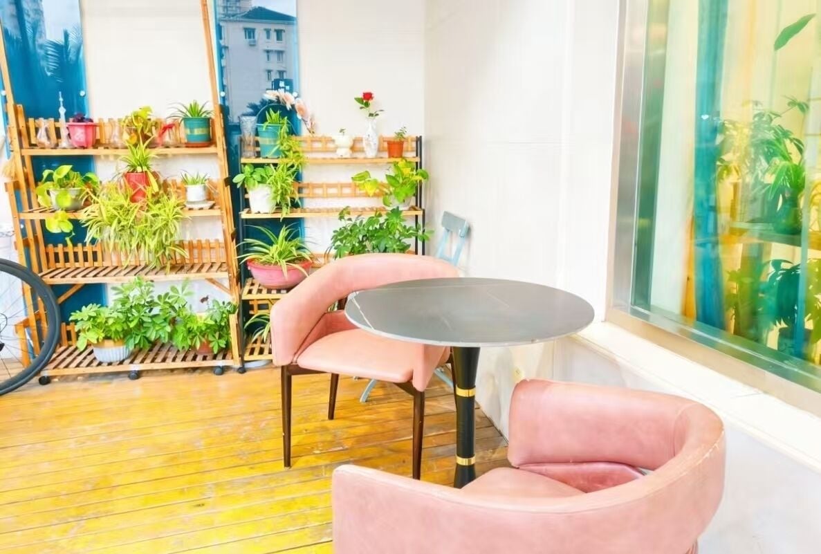 Een kamer met een ronde tafel, roze stoelen, een houten plantenplankje en uitzicht door een glazen raam.