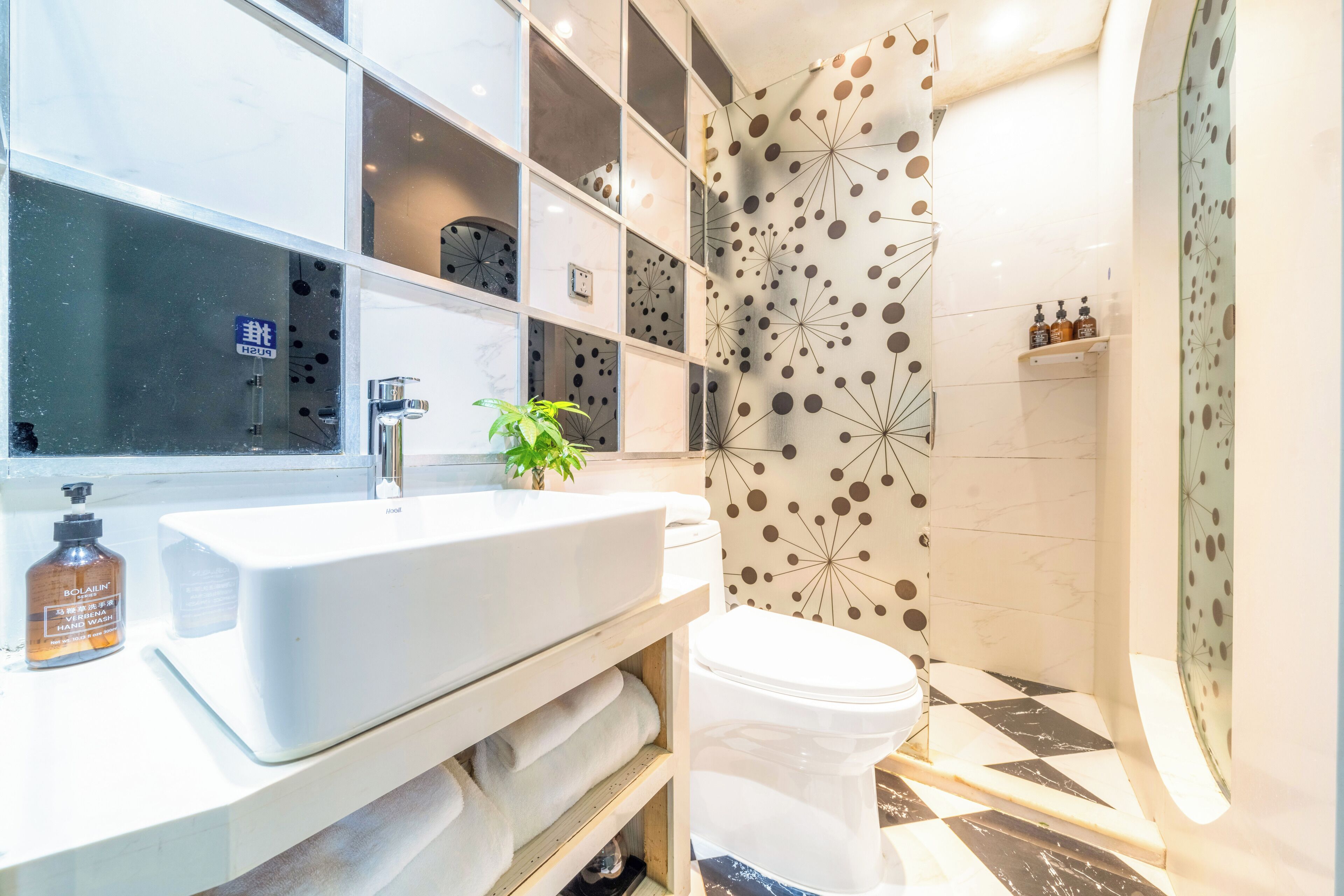 Une salle de bain moderne avec un lavabo blanc, des toilettes et un espace douche. On peut voir une plante en pot sur le comptoir et un flacon de savon liquide.