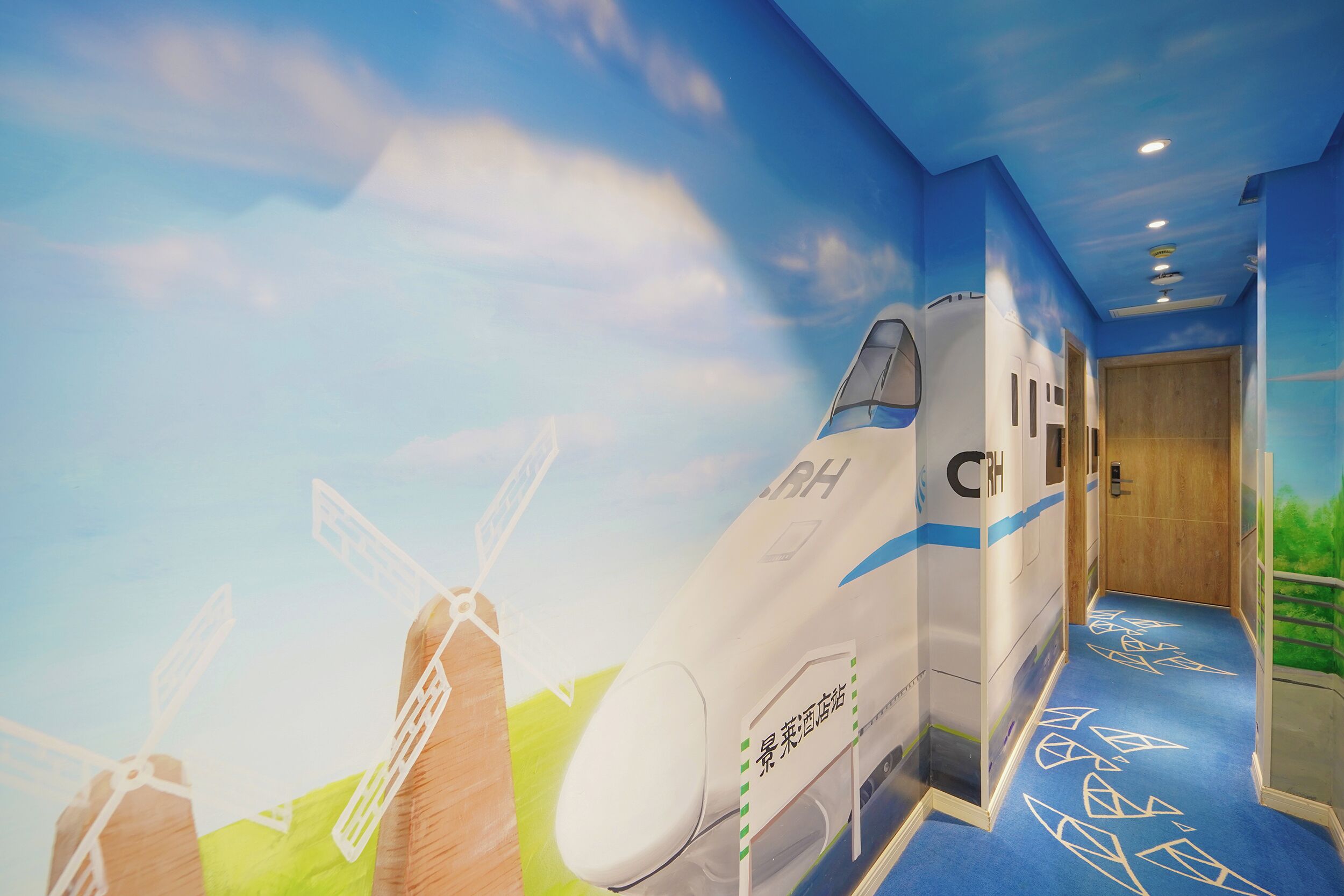 A corridor with a mural of a jet and windmills, a blue carpet with geometric patterns, and a wooden door at the end.