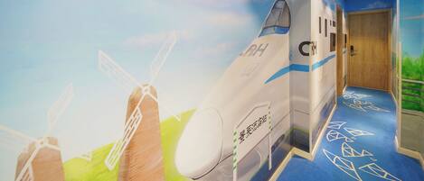 A corridor with a mural of a jet and windmills, a blue carpet with geometric patterns, and a wooden door at the end.