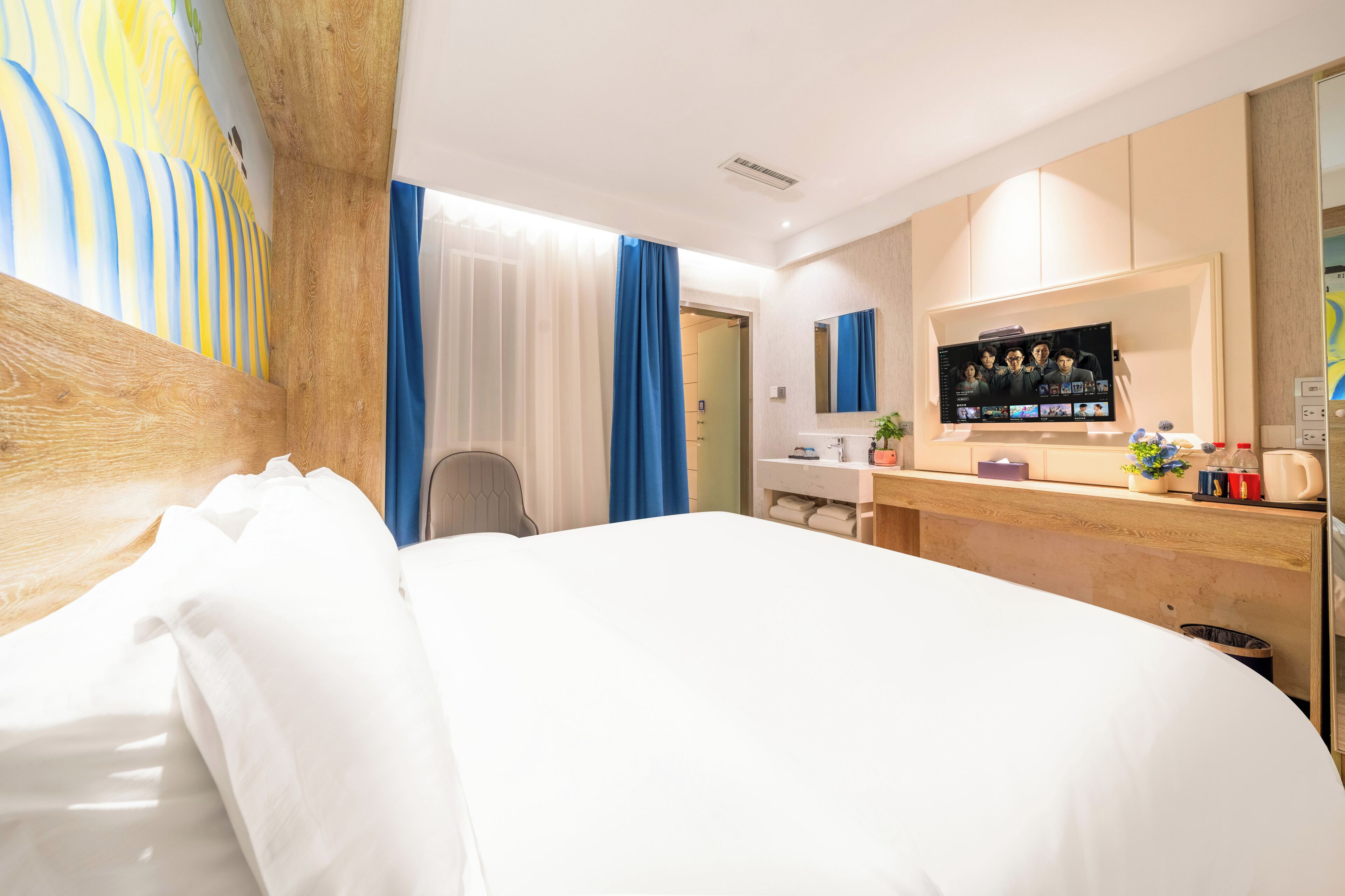 A hotel room with a large bed, a desk with a TV, a bathroom with a shower curtain, and a wall with a colorful mural.