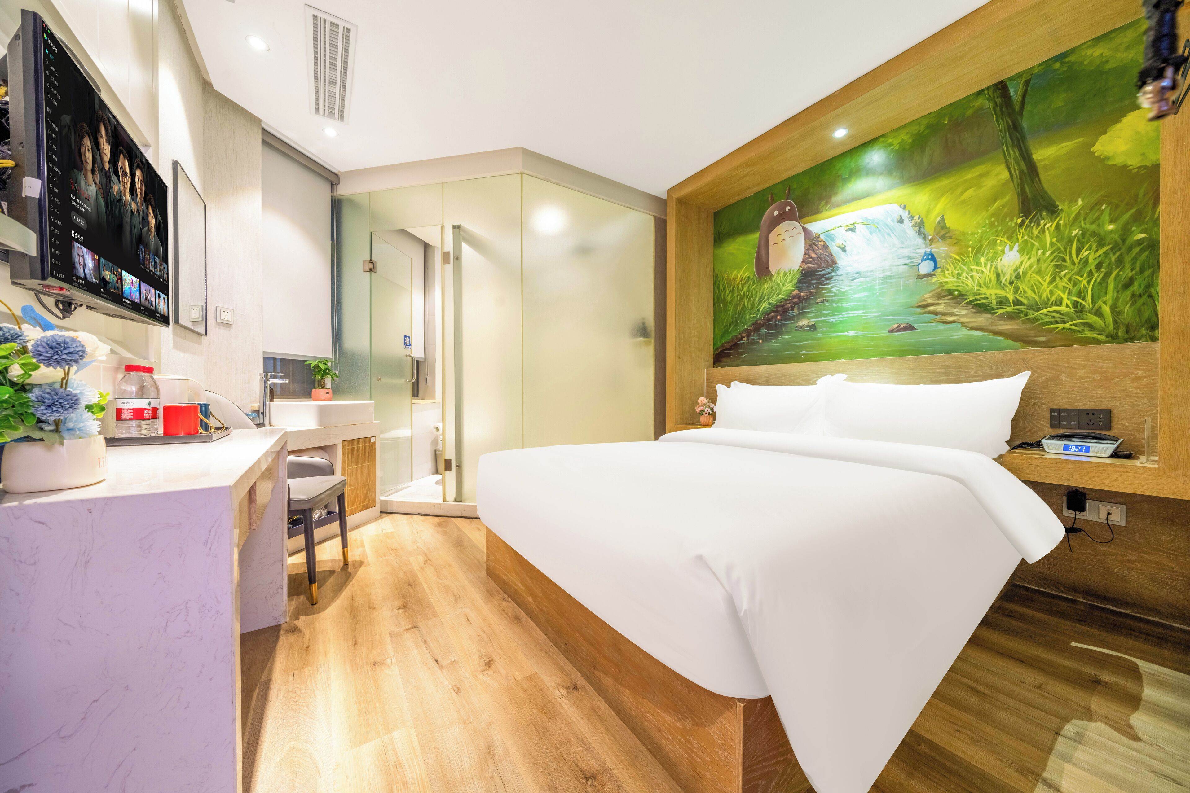 A modern hotel room with a wooden floor, a large bed, a desk with a computer, a TV mounted on the wall, and a mural of a forest scene.