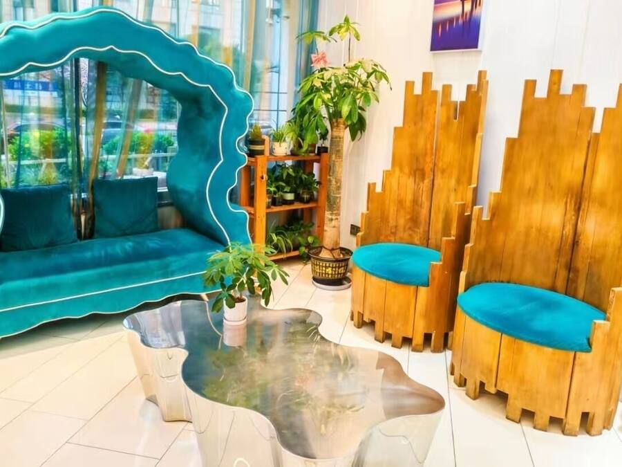 Un sofá color turquesa, sillas de madera, una mesa de centro de vidrio y una estantería con plantas.