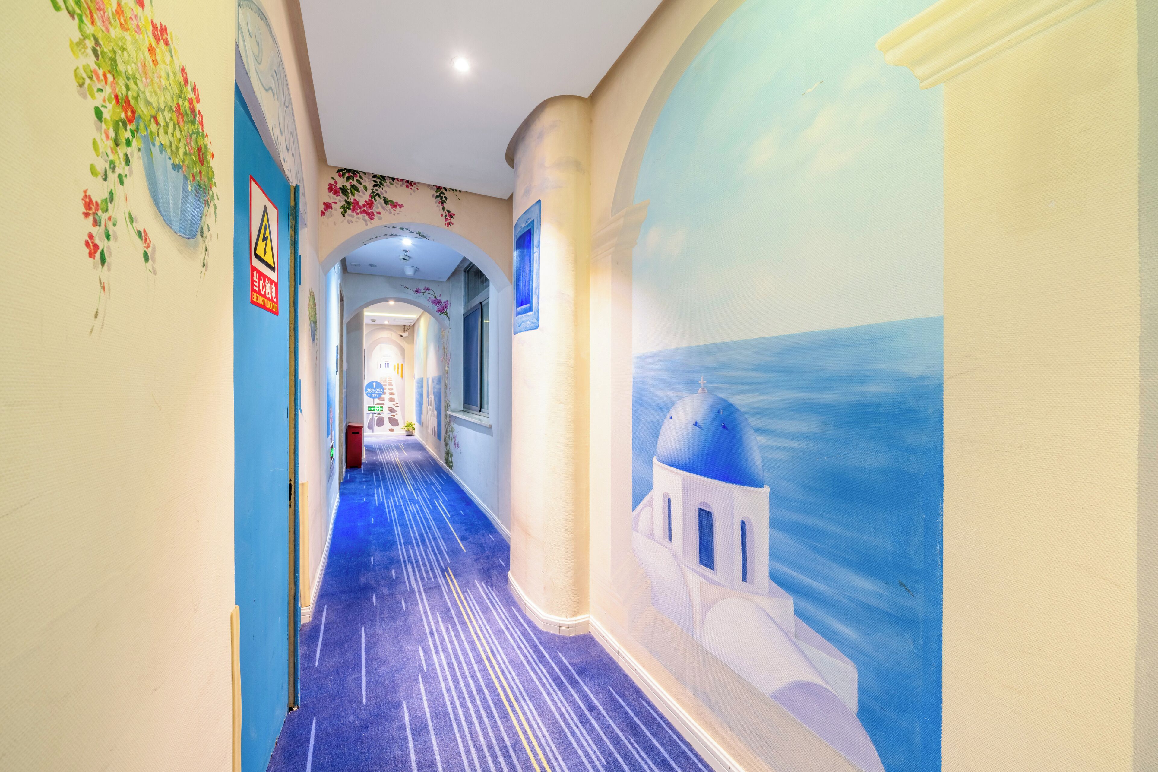 A hallway with a blue carpet, a mural of a blue sky and white building, and a warning sign on the wall.