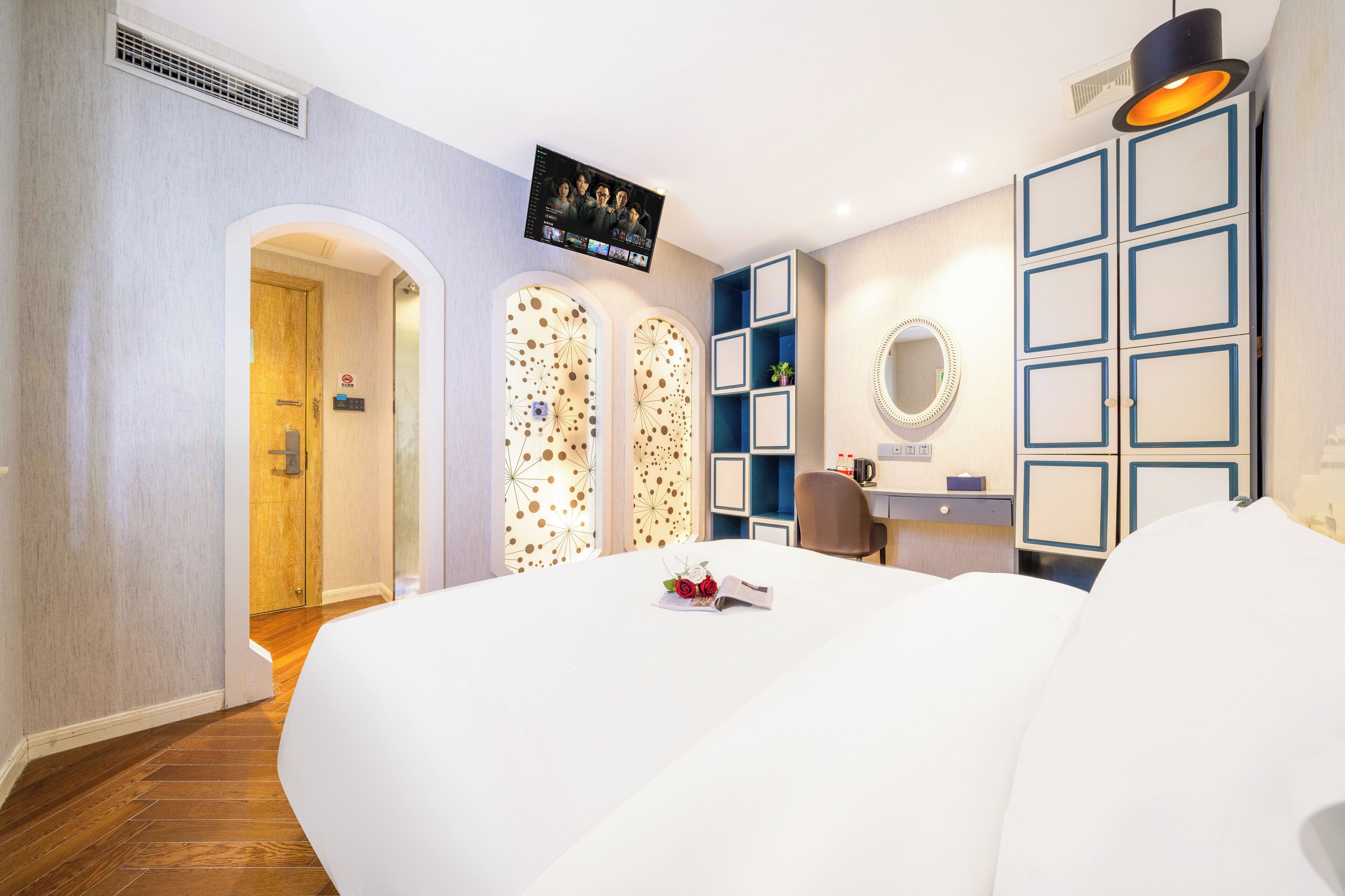 A modern hotel room with a large bed, a TV mounted on the wall, a desk with a chair, and a mirror.