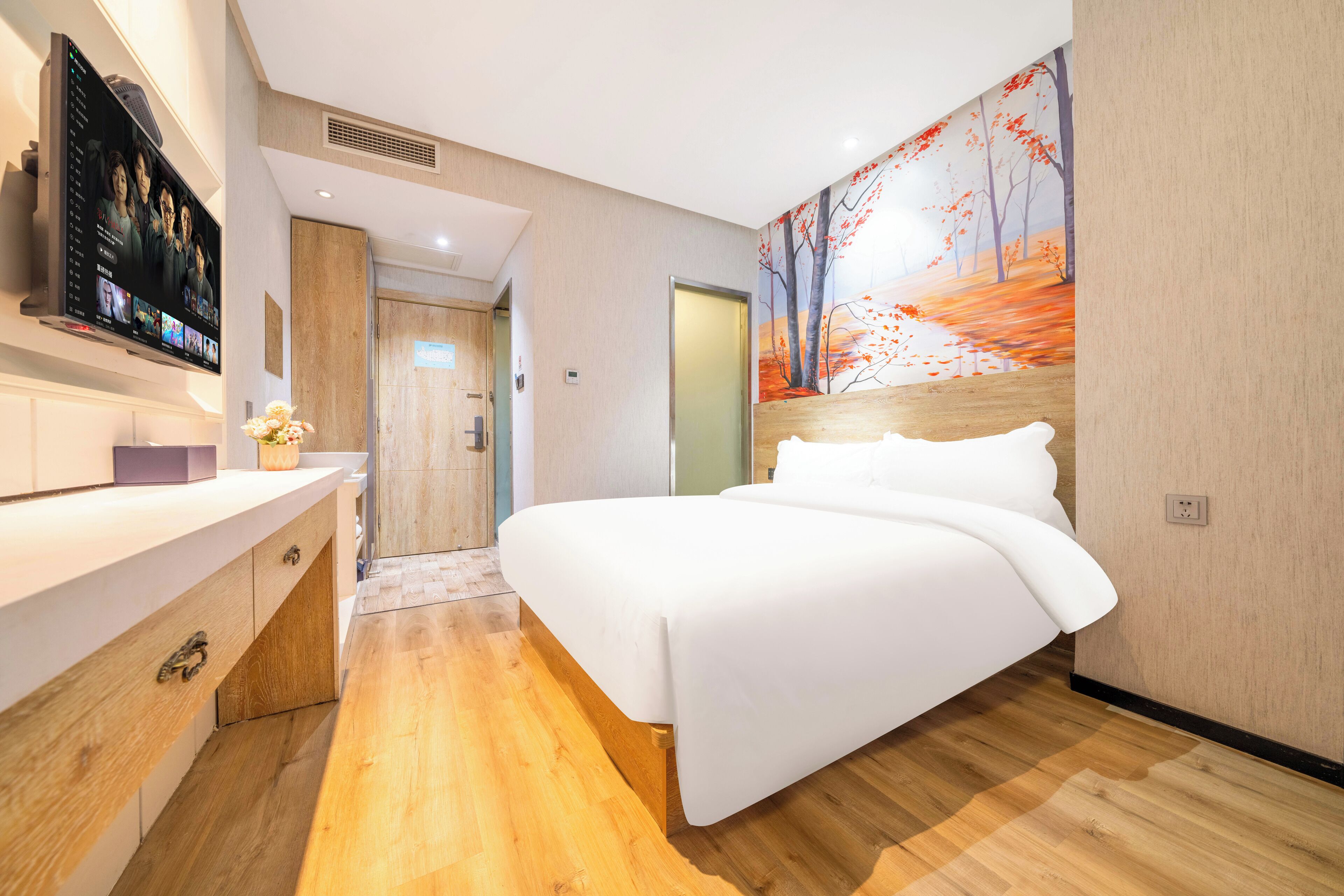 A modern hotel room with a wooden floor, a large bed with white linens, a flat-screen TV mounted on the wall, and a painting of autumn trees.