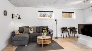 A modern living room with a grey sectional sofa, a round wooden coffee table, and a TV on a stand. There are two windows with views of trees, a floor lamp, and a wall-mounted lamp. - "Klint" - 400m from the sea by Interhome (Hals)