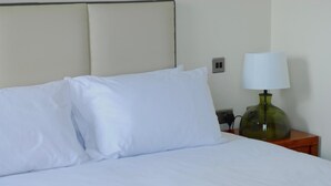 A neatly made bed with white linens, a green lamp on a wooden nightstand, and a wall socket. - Icon Hotel  (Luton)