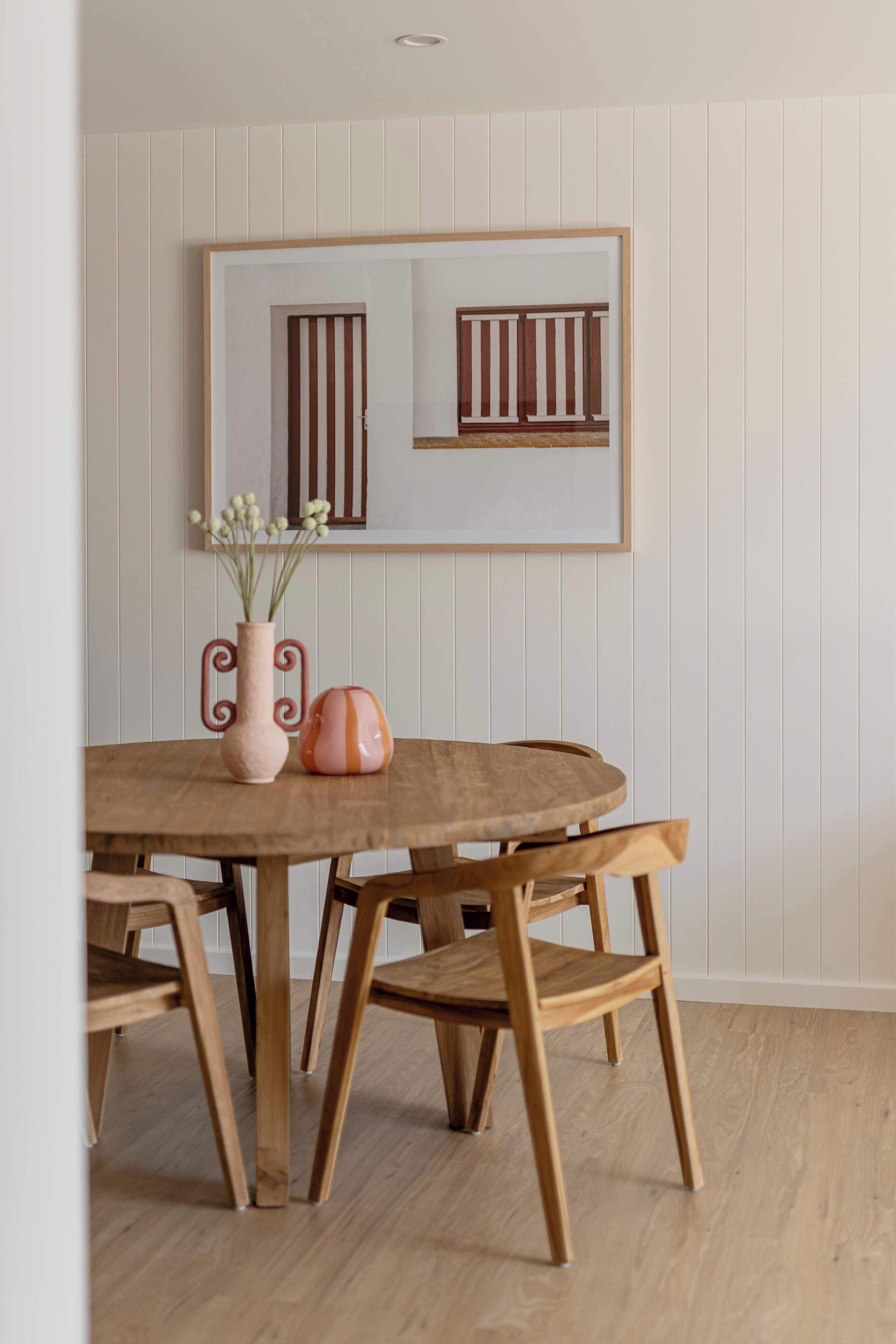 A dining area with a wooden table and chairs, a vase with flowers, and a framed picture on the wall.