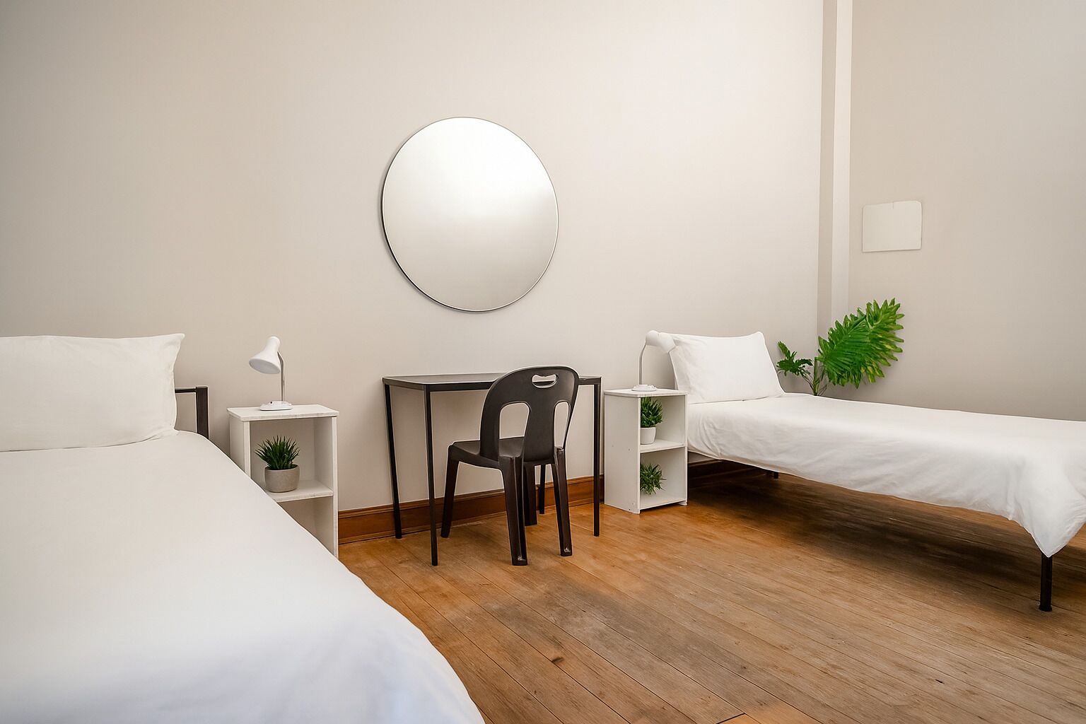 Ein Hotelzimmer mit zwei Betten, einem Schreibtisch mit Stuhl, einem runden Spiegel an der Wand und einer kleinen Pflanze.