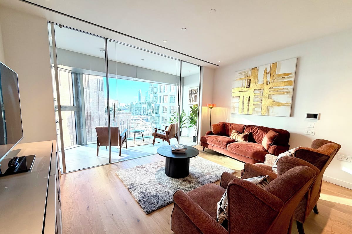 A modern living room with a large glass door leading to a balcony, a brown sofa set, a coffee table, and a large abstract painting on the wall.
