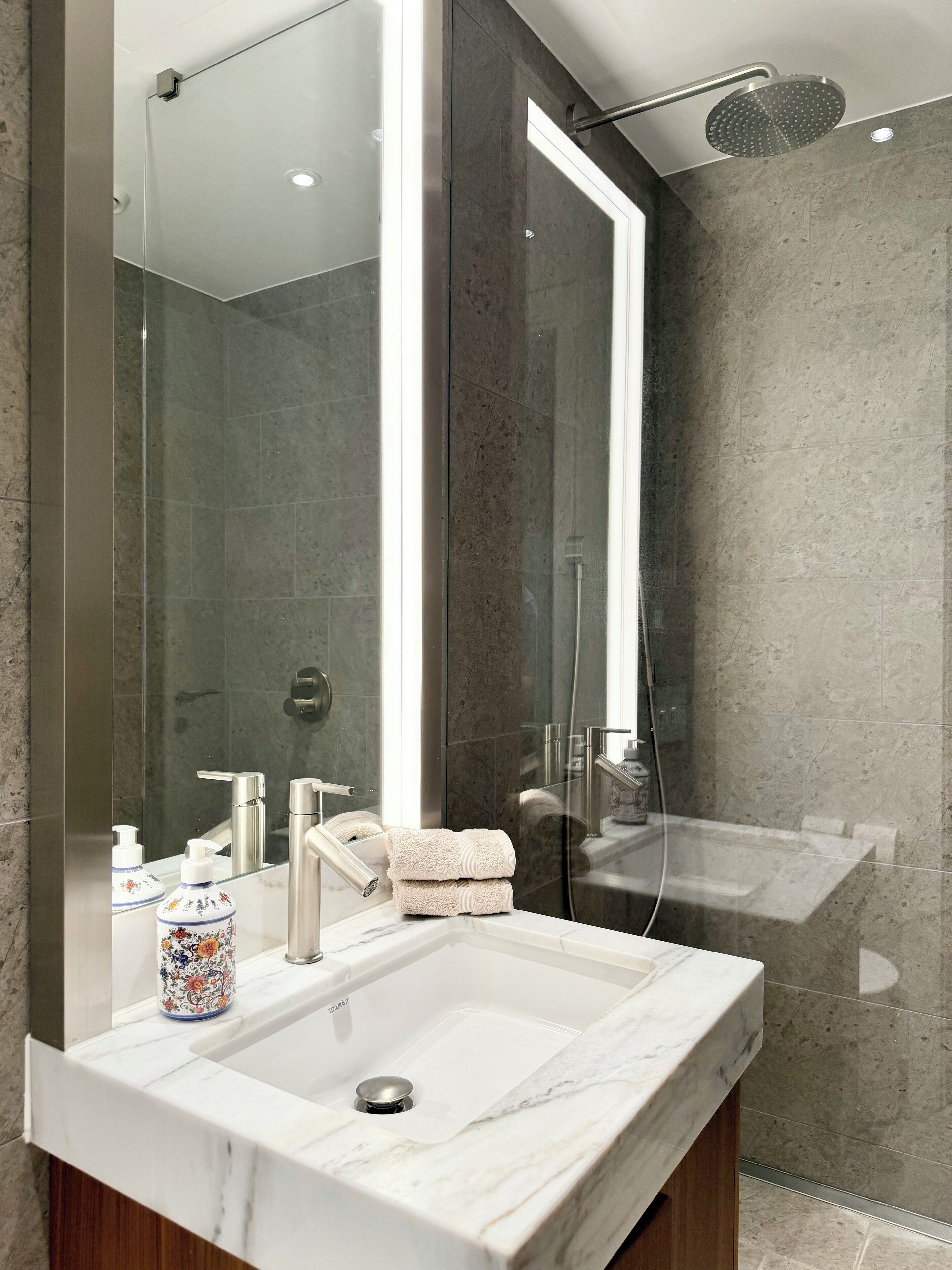 A modern bathroom with a large mirror, a marble sink, a glass shower enclosure, and a towel on the counter.