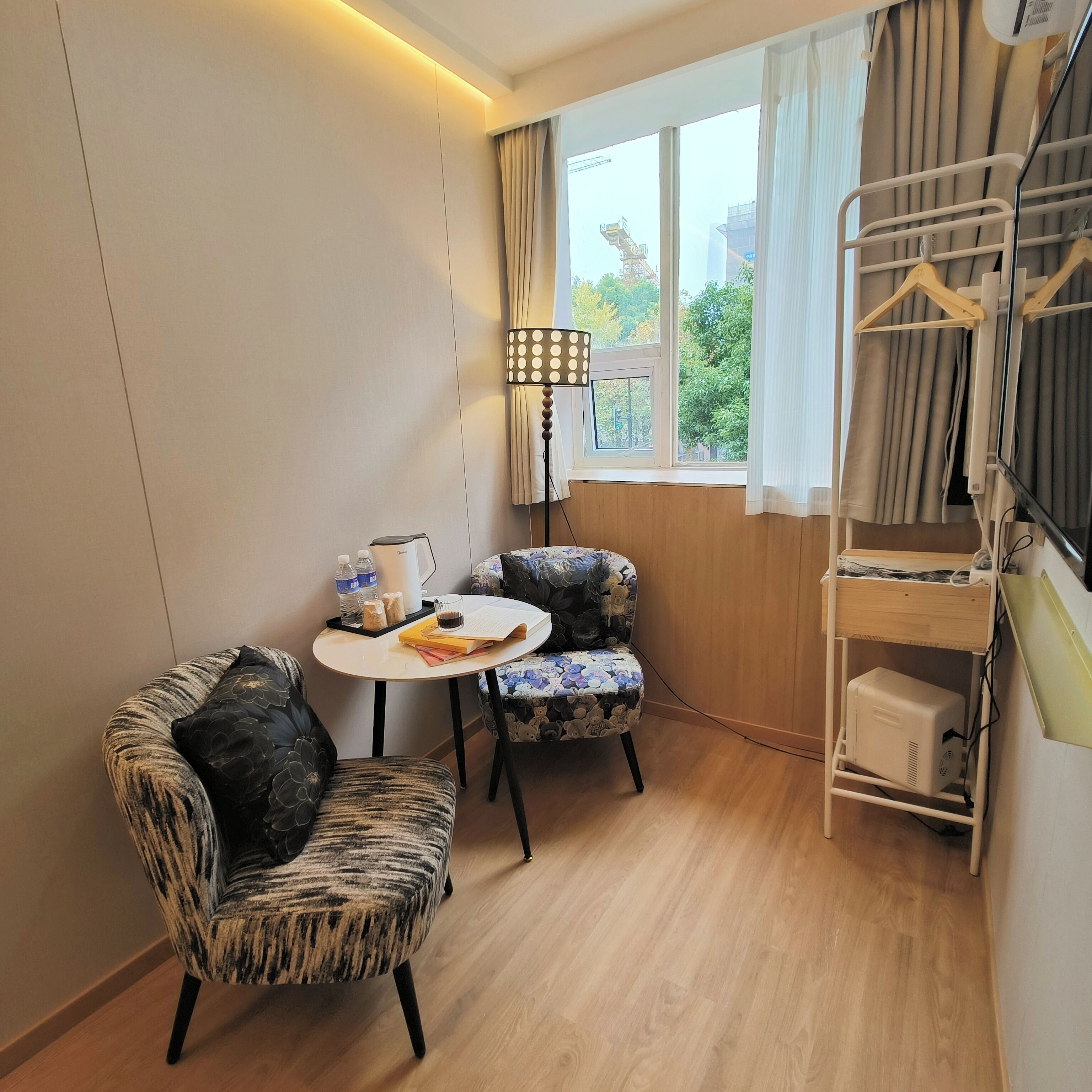 A small room with two patterned armchairs, a small table with a lamp, a window with curtains, and a clothing rack.