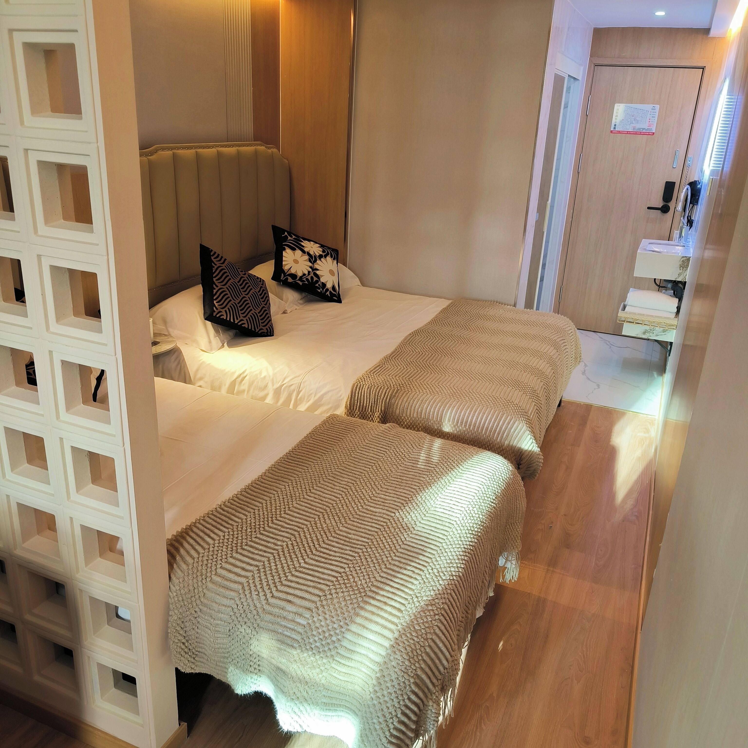 Habitación de hotel con dos camas, piso de madera, separador blanco con estantes abiertos y baño con lavabo y perchero para toallas.