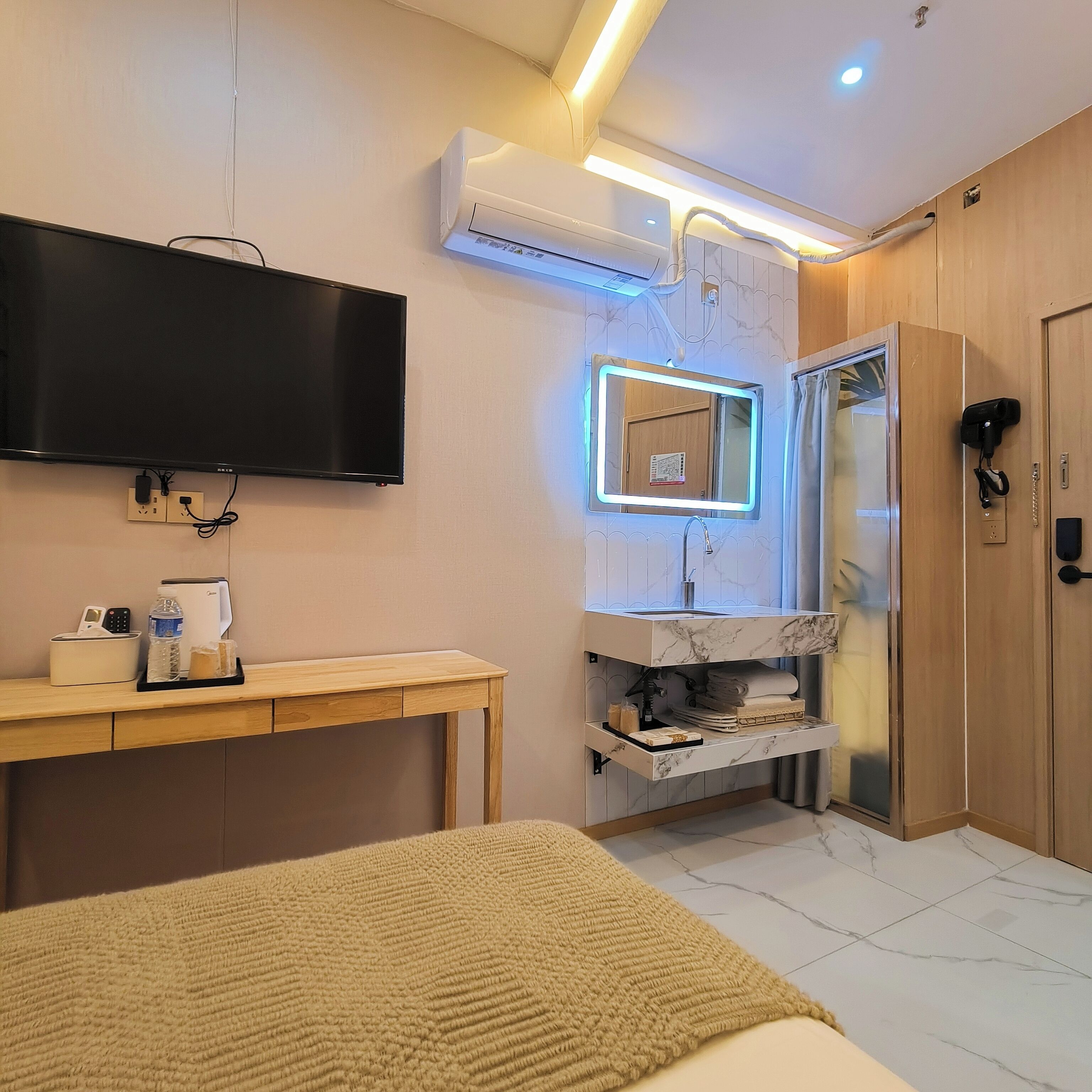 A modern hotel room with a flat-screen TV, a built-in air conditioner, a vanity with a mirror, a wooden desk, and a bed.
