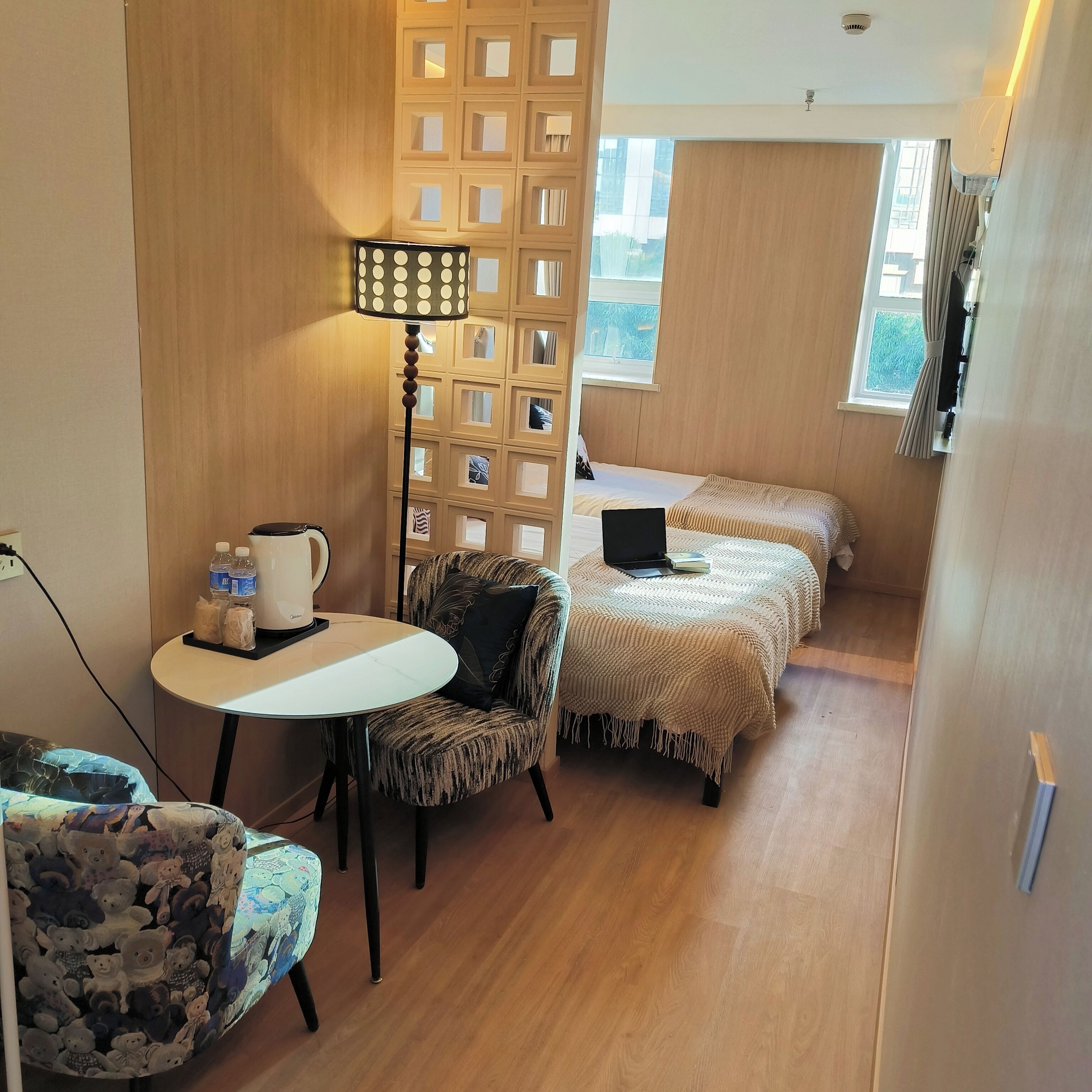 A compact hotel room with a wooden floor, a round table with a lamp, a chair, two beds with blankets, and a window with curtains.