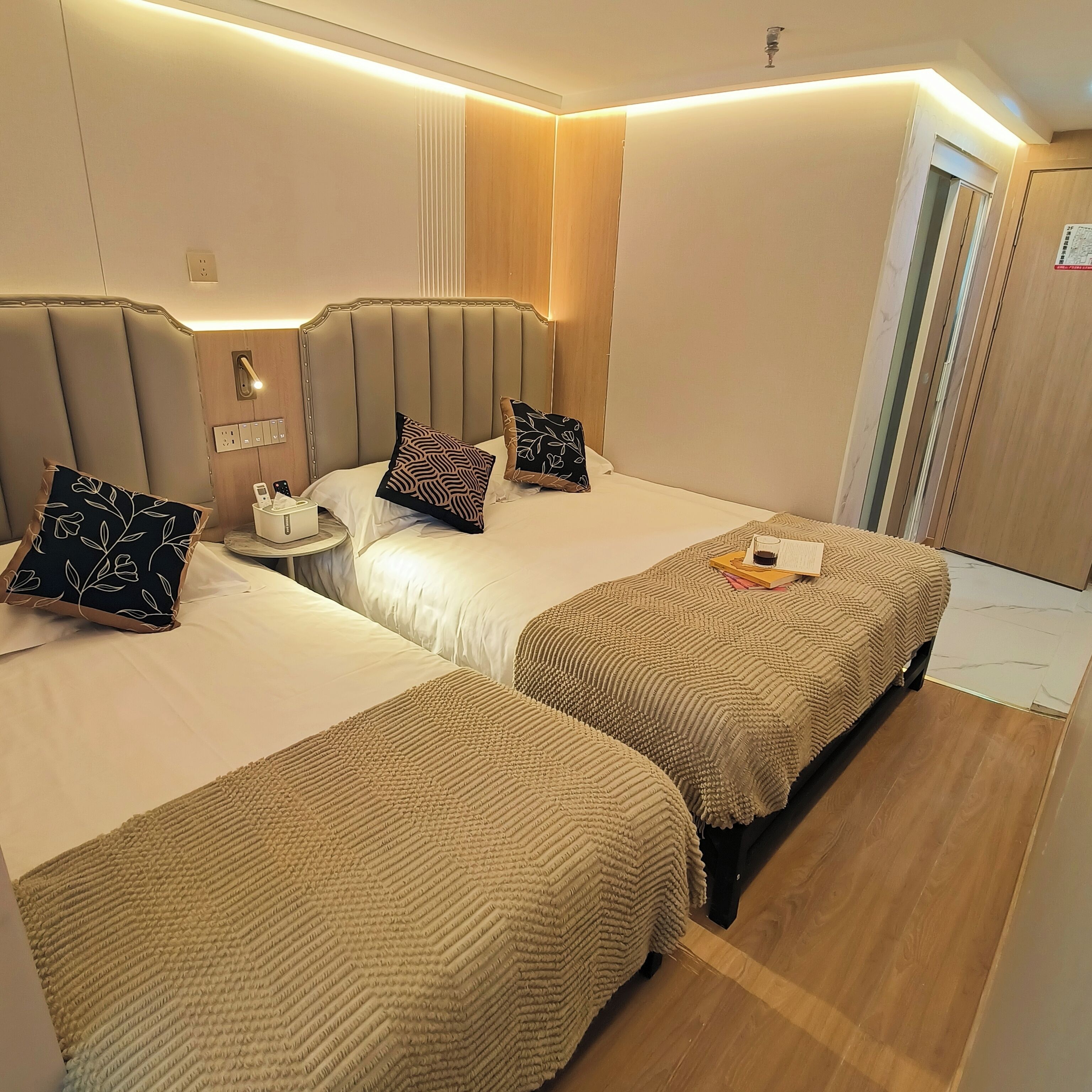 A hotel room with two beds, a bedside table with a lamp, and a small table with a phone and tissues.
