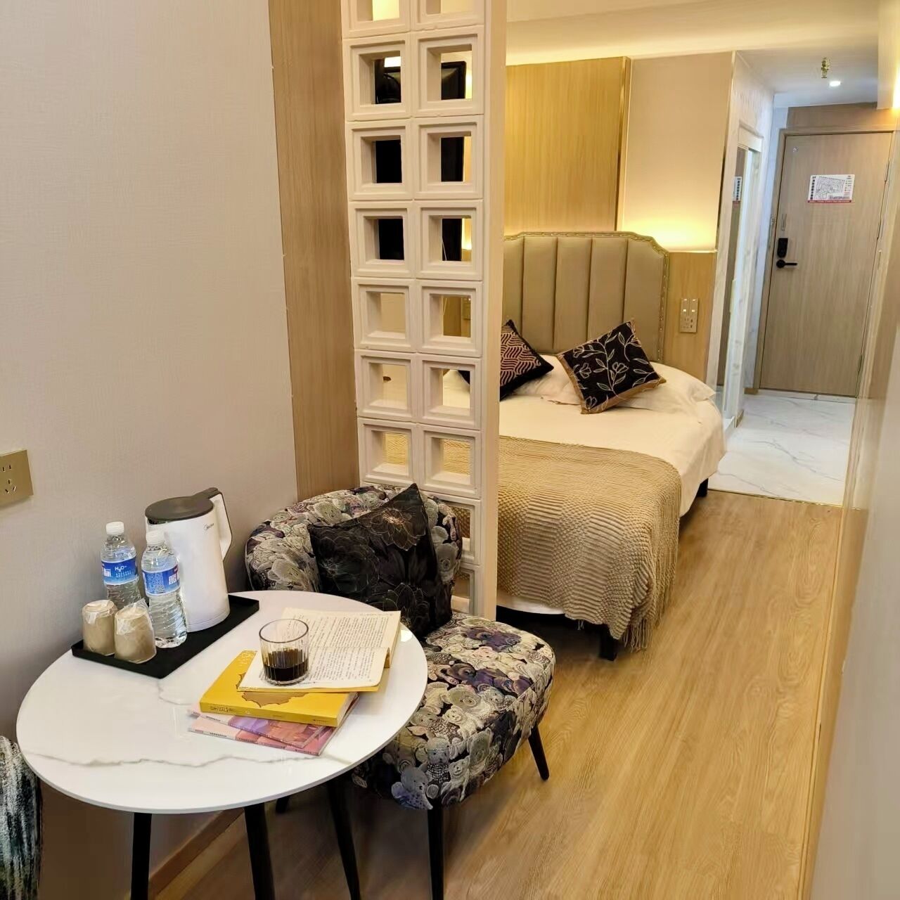 Habitación de hotel con una cama, una mesita con libros y un vaso, una silla decorativa y un separador con diseños geométricos.