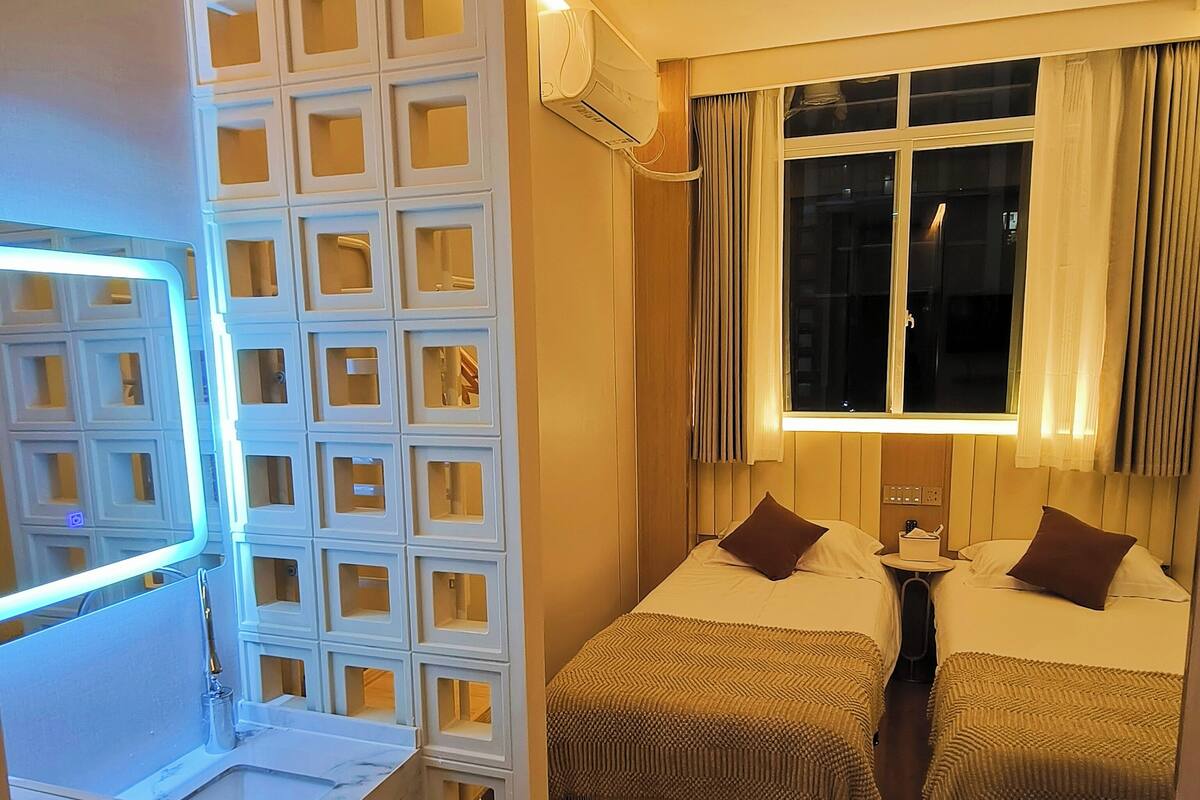 A hotel room with two beds, a desk with a lamp, a window with curtains, and a wall-mounted air conditioner.