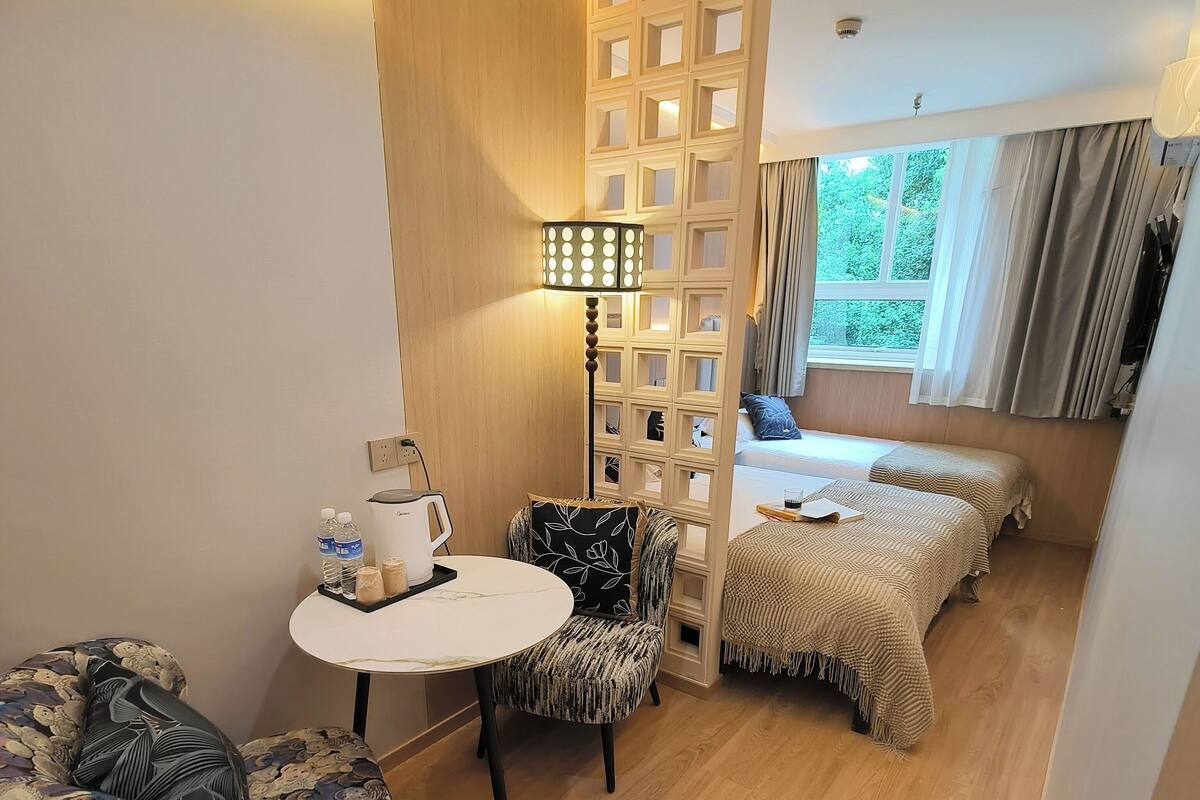 A compact hotel room with a bed, a small table with a kettle, a chair, and a decorative partition.