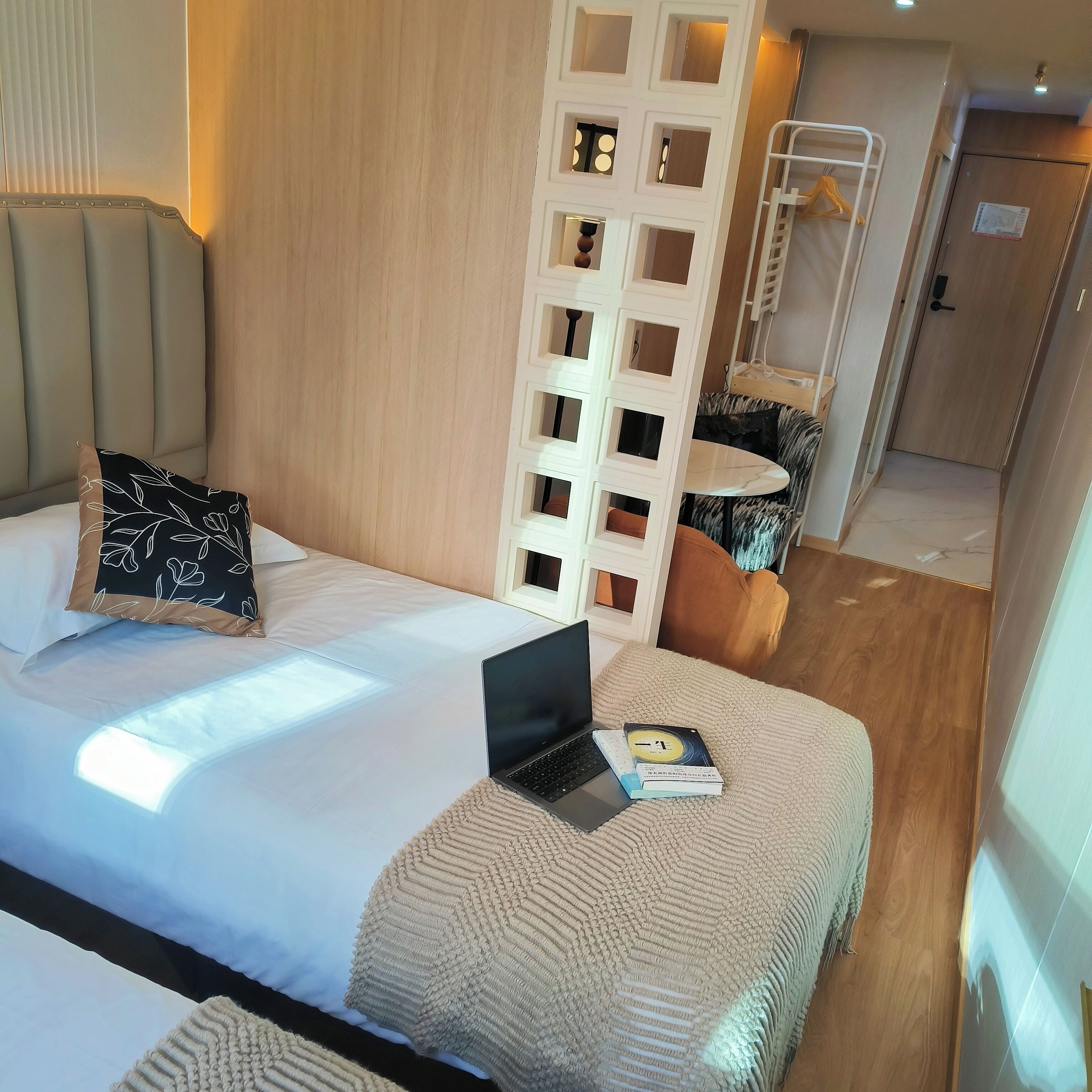 Une chambre d’hôtel avec un lit, un ordinateur portable et un livre posé sur le lit. On peut voir un plancher en bois, un mur blanc et une porte au fond.