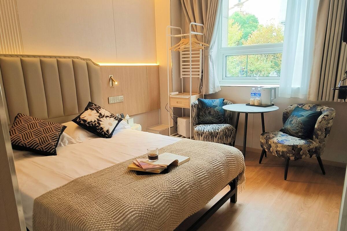 A hotel room with a large bed, a small round table, two chairs, a window with curtains, and a wall-mounted lamp.