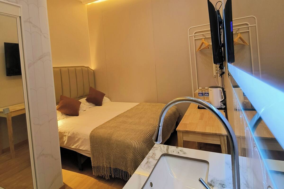 A hotel room with a bed, a bedside table with a kettle, a mirror, and a sink.