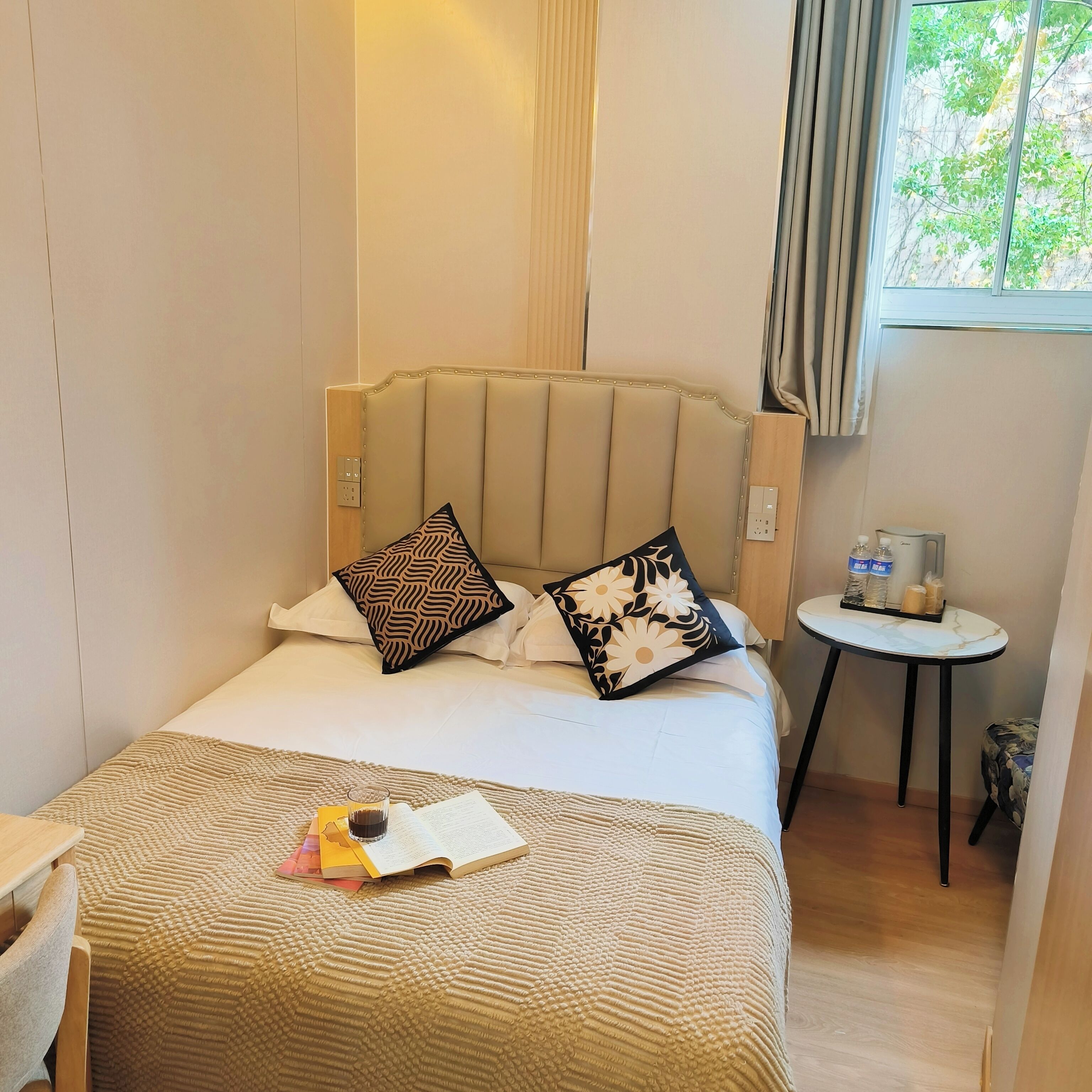 A hotel room with a bed, a small table with a water bottle and a candle, and a window with curtains.