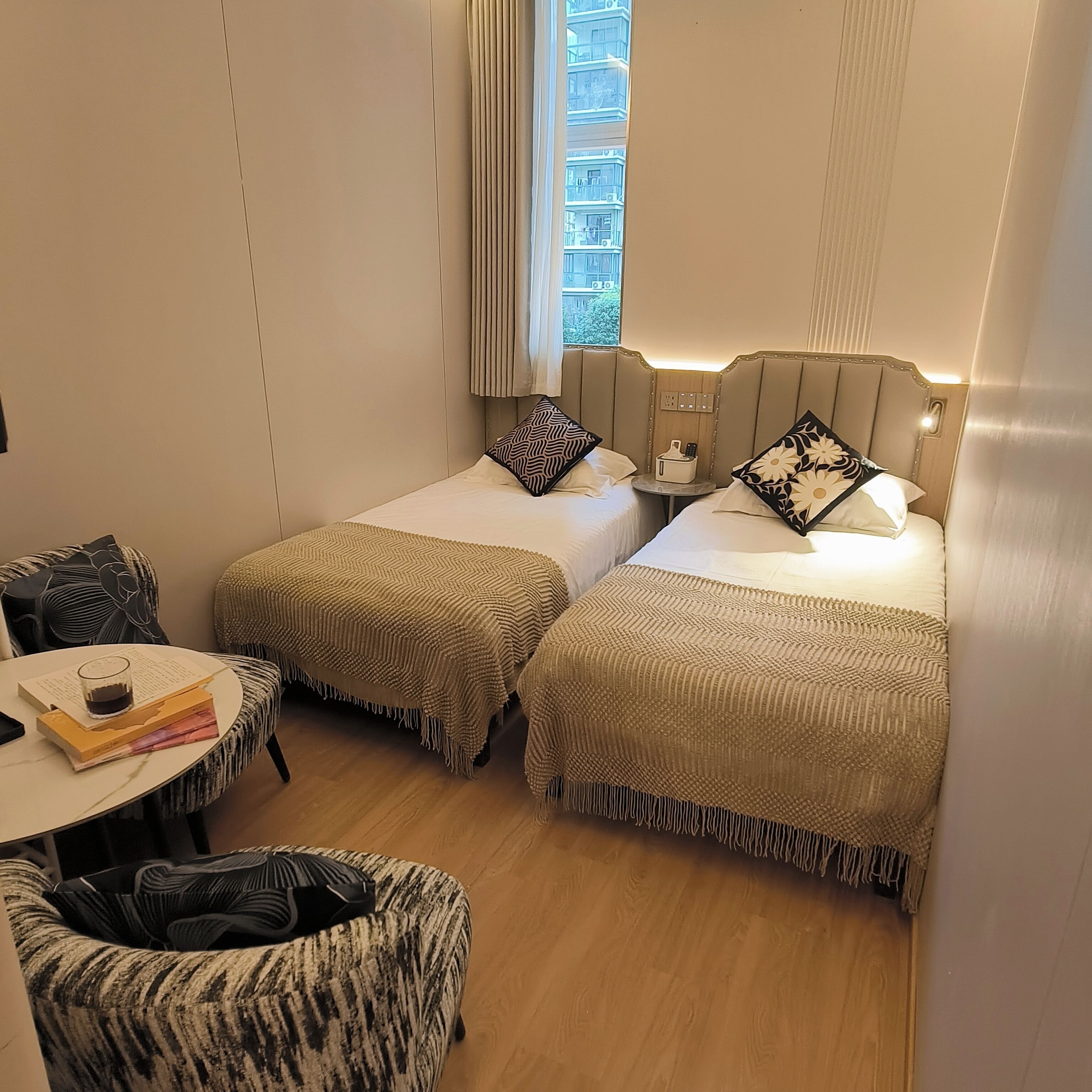 A hotel room with two beds, a small table with a chair, and a window with curtains.