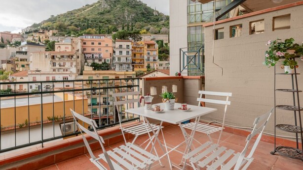 Een balkon met witte stoelen en een tafel, met uitzicht op een stadsgezicht met gebouwen en een heuvel.