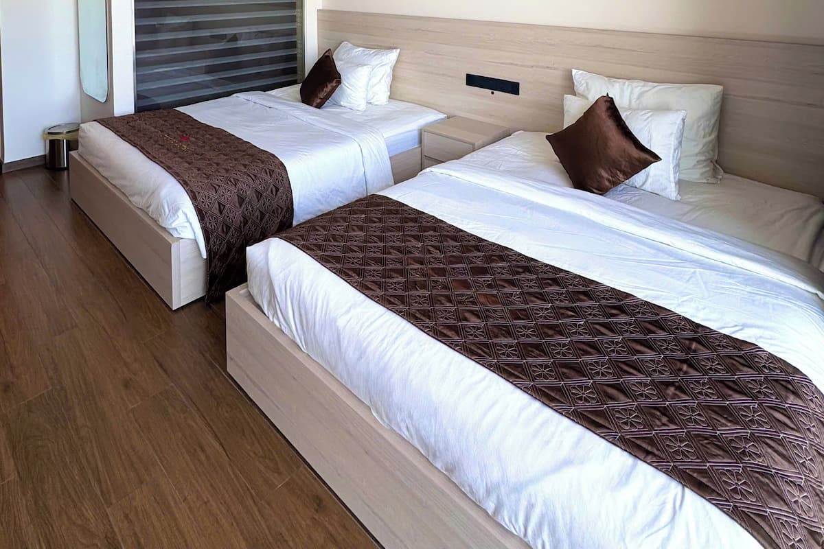 A hotel room with two beds, a large window with blinds, a bedside table, and a small trash bin.
