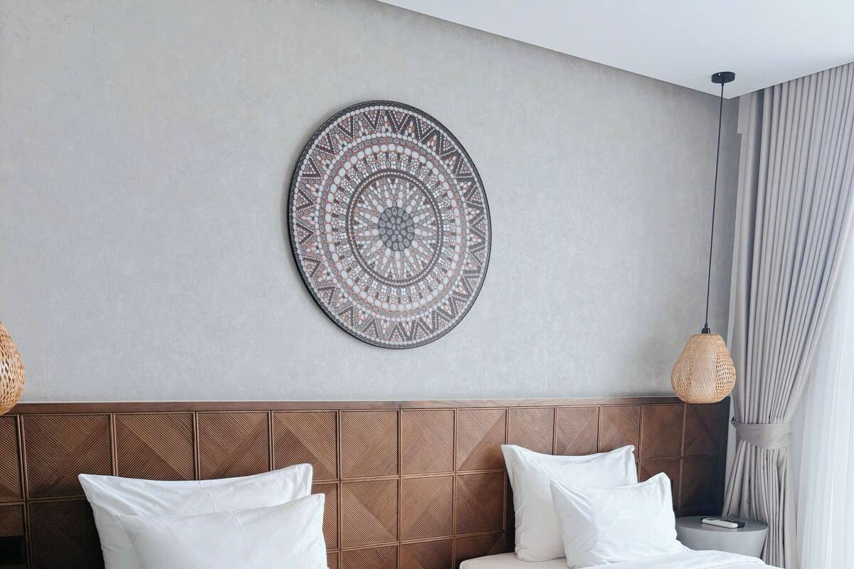 A hotel room with two beds, a decorative wall piece, a woven light fixture, and a bedside table with a remote control.
