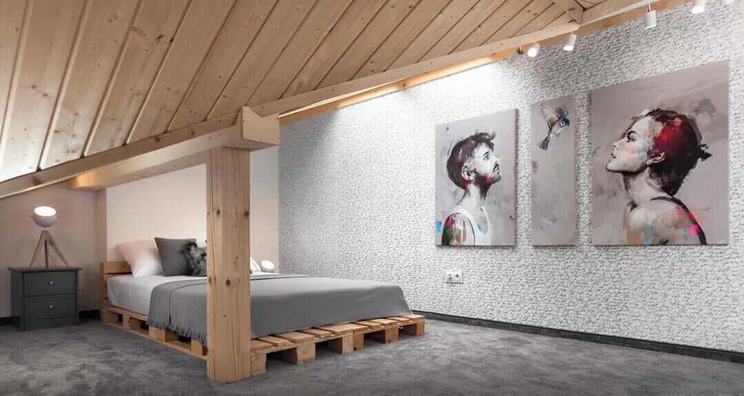 Een slaapkamer met een houten plafond, een bed met een grijze deken, een grijze nachtkastje en twee aan de muur hangende schilderijen.