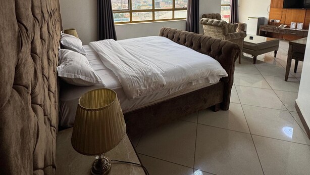 A hotel room with a large bed, a desk, a chair, a TV, and a window with curtains. - Sanso Hotel (Kampala)