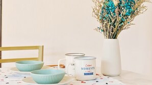 Una mesa de comedor con mantel blanco, tres cuencos azul claro y un jarrón blanco con flores secas.