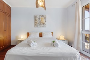 Una cama bien tendida con dos toallas enrolladas, un cabecero de madera, una obra de arte en la pared y una lámpara de noche.