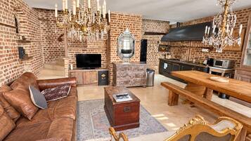 A spacious living room with a leather sofa, a wooden coffee table, a dining table with benches, a chandelier, and a brick wall.