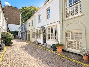 A cobblestone street lined with white buildings, some with arched windows and doors, and potted plants along the sidewalk. - Knightsbridge 2BR Mews House (London)