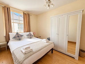 Un dormitorio con cama, almohadas, mesita de noche, lámpara, armario y ventana con cortinas.
