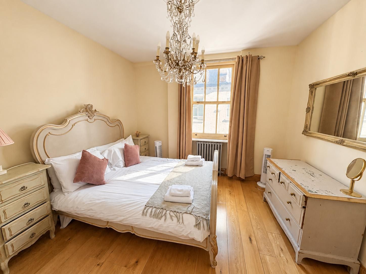 A bedroom with a large bed, a dresser, a mirror, a chandelier, and a window with curtains.