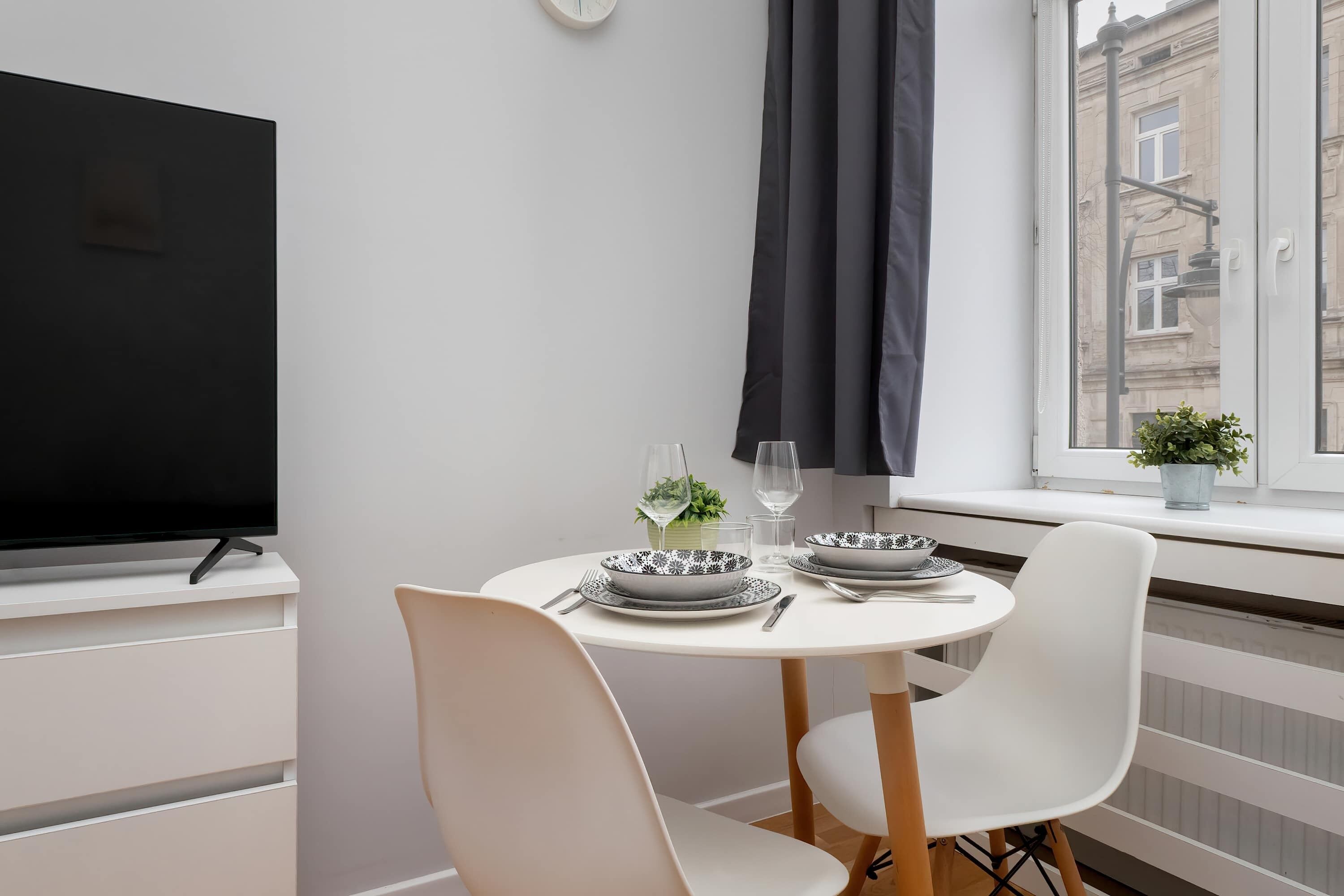 Un comedor con una mesa redonda preparada para dos personas, un televisor sobre un mueble blanco y una ventana que ofrece vistas a edificios.