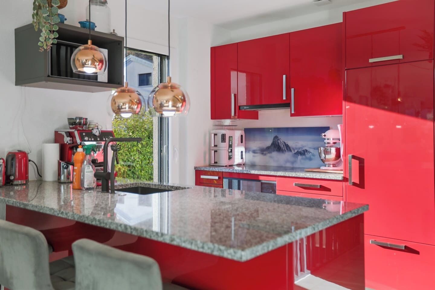 A modern kitchen with red cabinets, a granite countertop, and pendant lighting.