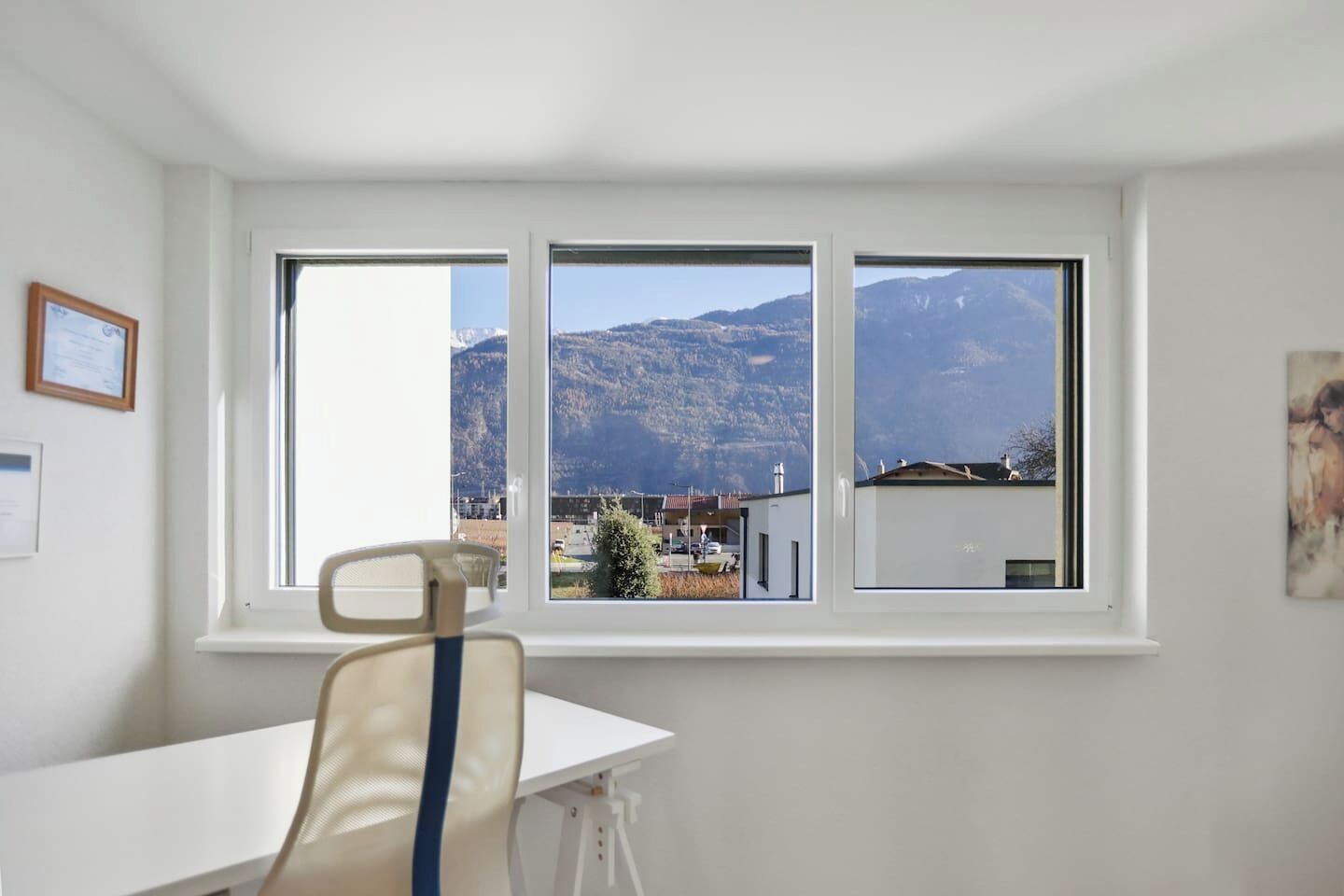 Quarto com uma grande janela que oferece vista para as montanhas, uma mesa branca e uma cadeira.