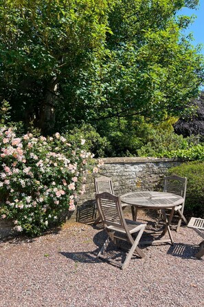Ein Garten mit einer Steinsäule, einem Tisch mit vier Stühlen und blühenden rosa Blumen.