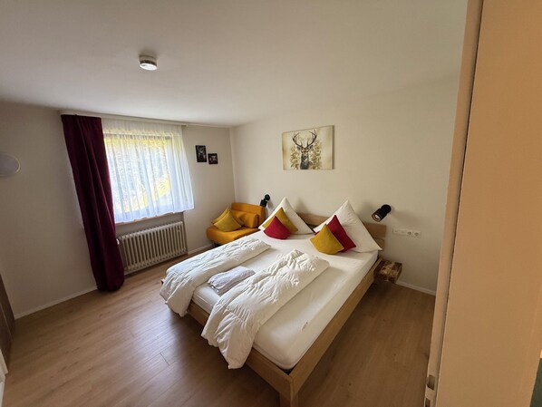 A bedroom with a large bed, a chair, a radiator, a window with curtains, and a wall-mounted light. - House Anna Alpirsbach (Alpirsbach)