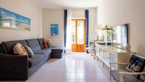 Ein Wohnzimmer mit einer Couch, einem Fernseher und einem Sessel. Eine Tür führt zu einem Balkon.