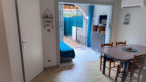 Et værelse med et spisebord, stole, et fjernsyn og en dør, der fører ud til en balkon med et blåt gardin.