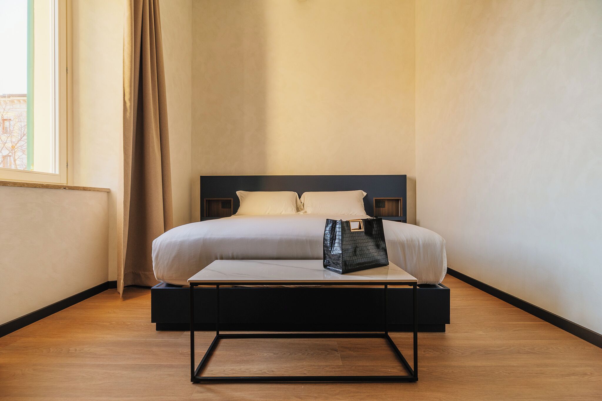 A neatly made bed with a black metal frame, a white mattress, and two pillows. A black handbag is placed on the bed. The room has wooden flooring and a window with curtains.