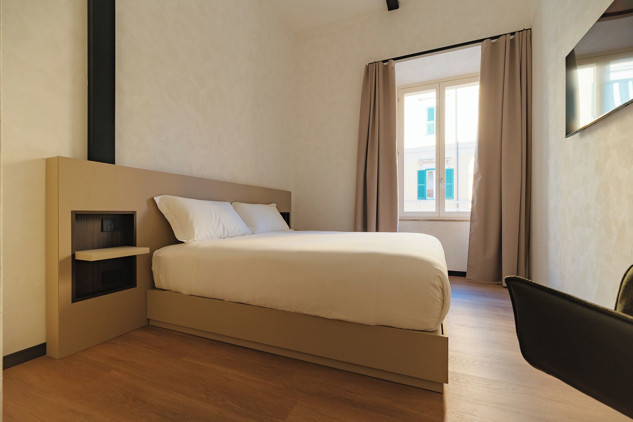 A modern hotel room with a bed, a small bedside table, a chair, and a window with curtains.